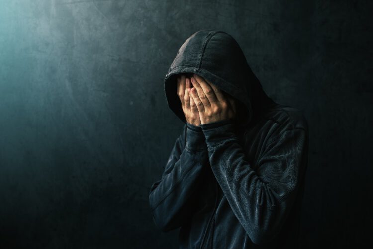 Desperate man in hooded jacket is crying, hands are covering face and tears in the eyes, light of hope shining from his right side