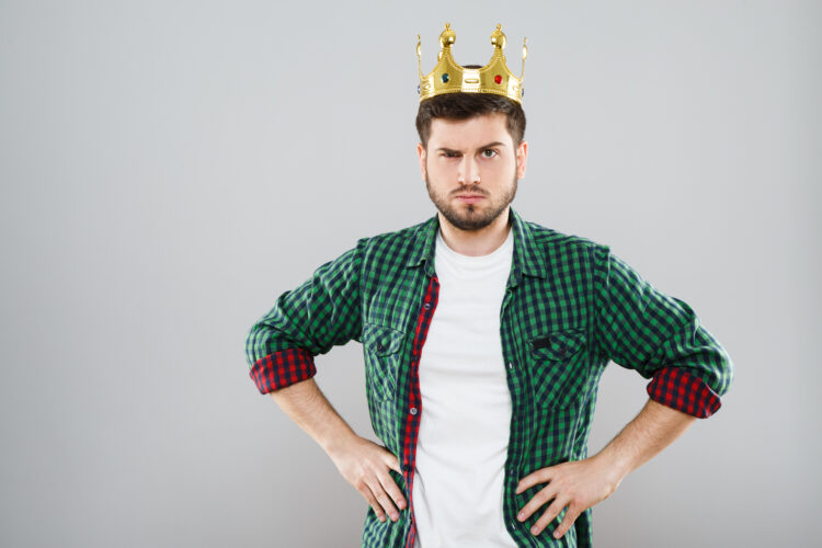 Man in green checked T-shirt and crown on his head. Looking at camera. Young man, clothing, casual style. Serious, frowning. Waist up. Indoors, studio