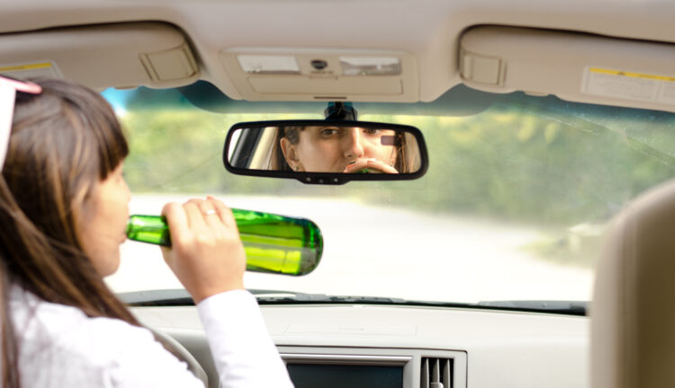 View from behind inside a car of a woman driver drink driving lifting the bottle to her lips and gulping down the alcohol as she steers the car
