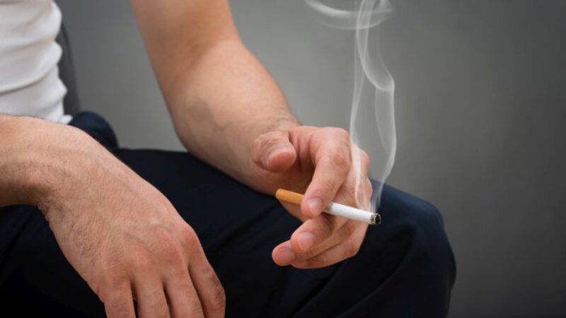 Midsection of man holding cigarette while sitting against gray background