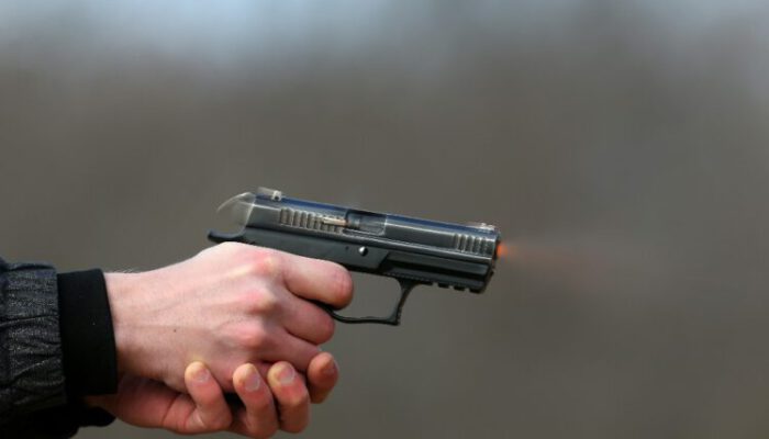 Close-up Photo of a pistol shooting with two hands, the shells emanating from the shutter and blue smoke on a blurred background.